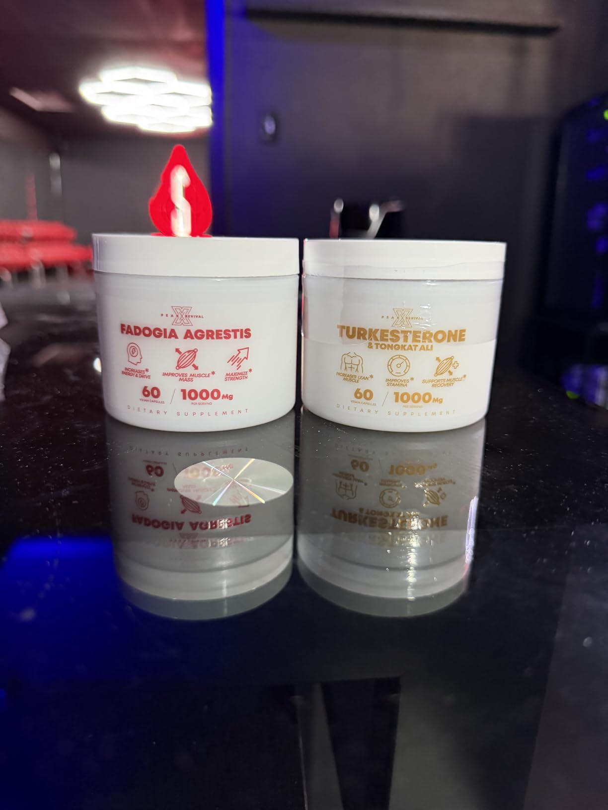 Two white jars, Fadogia Agrestis and Turkesterone and Tongkat Ali, 60 capsules and 1000 mg each, on a glossy black gym counter